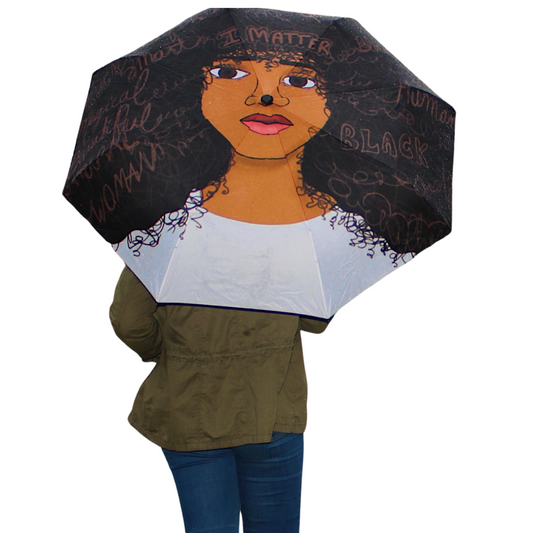 I Matter Umbrella that features deep brown and black accents and a white  lower area. The umbrella features a woman with words across her forehead that says "I Matter". Words featured in each strand of her hair like "beautiful", "queen", "human", "unique".  Black polyester umbrella featuring “I Matter” affirmation text, Afrocentric artwork, semi-automatic open canopy. 